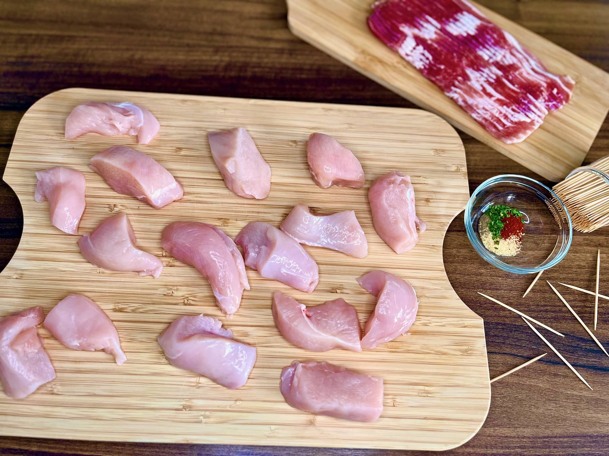 Chunks of chicken breast rest on a chopping board, accompanied by bacon strips and toothpick skewers