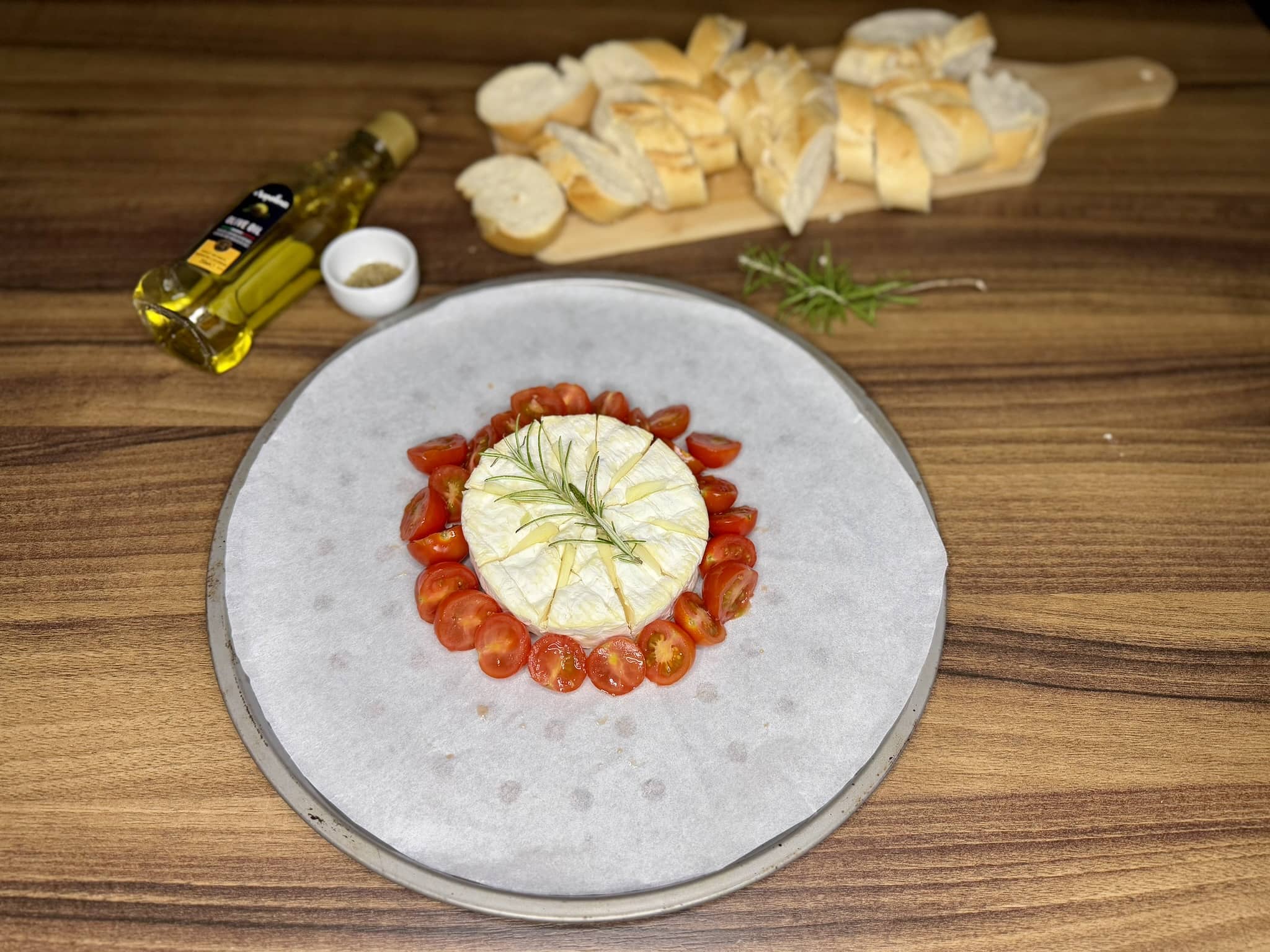 Camembert arranged in the centre of a tray, surrounded by halved tomatoes with rosemary on top of the cheese