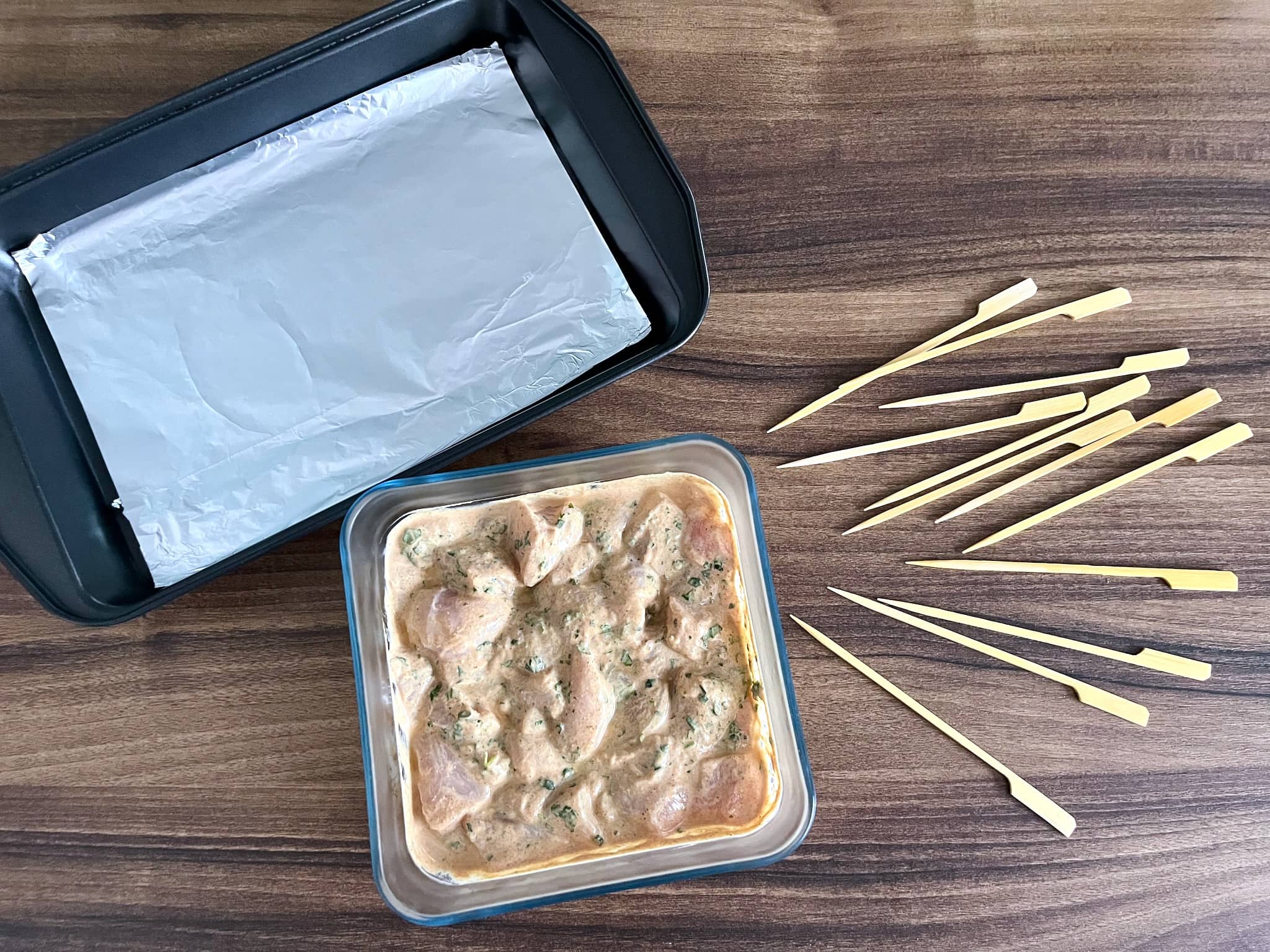 Marinaded chicken in a bowl, baking sheet lined with aluminium foil, and skewers on the side