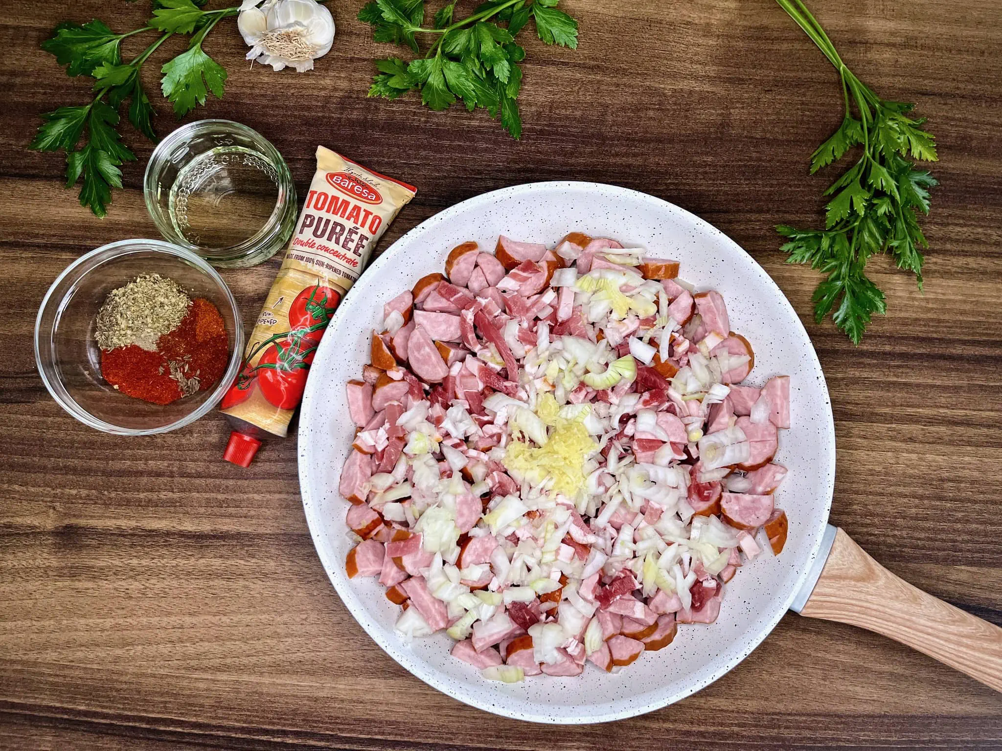 Pokrojone kiełbasy, boczek i inne składniki na patelni, z dodatkowymi przyprawami obok