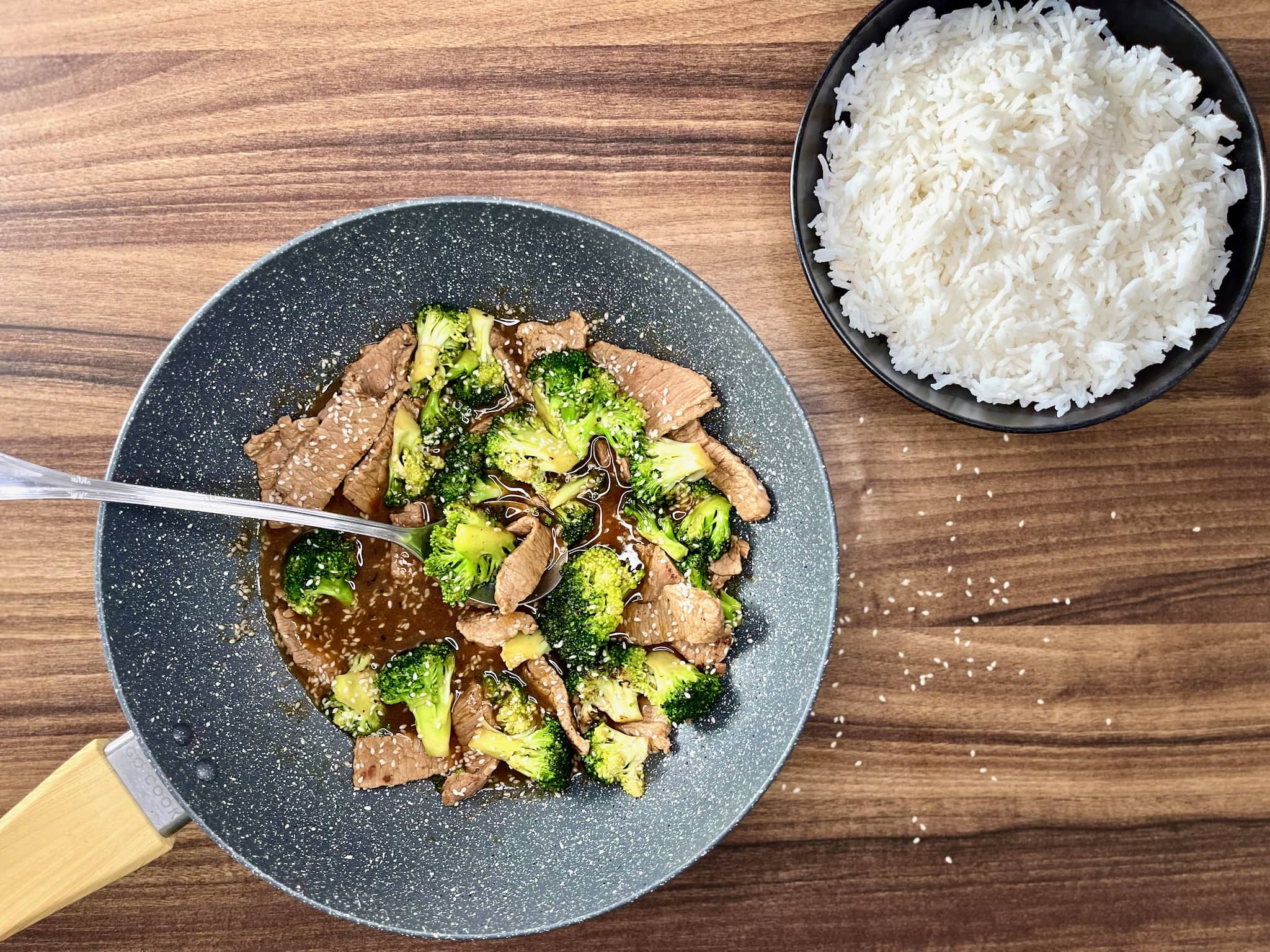 Beef and broccoli with egg noodles in a pan, with a bowl of rice on the side