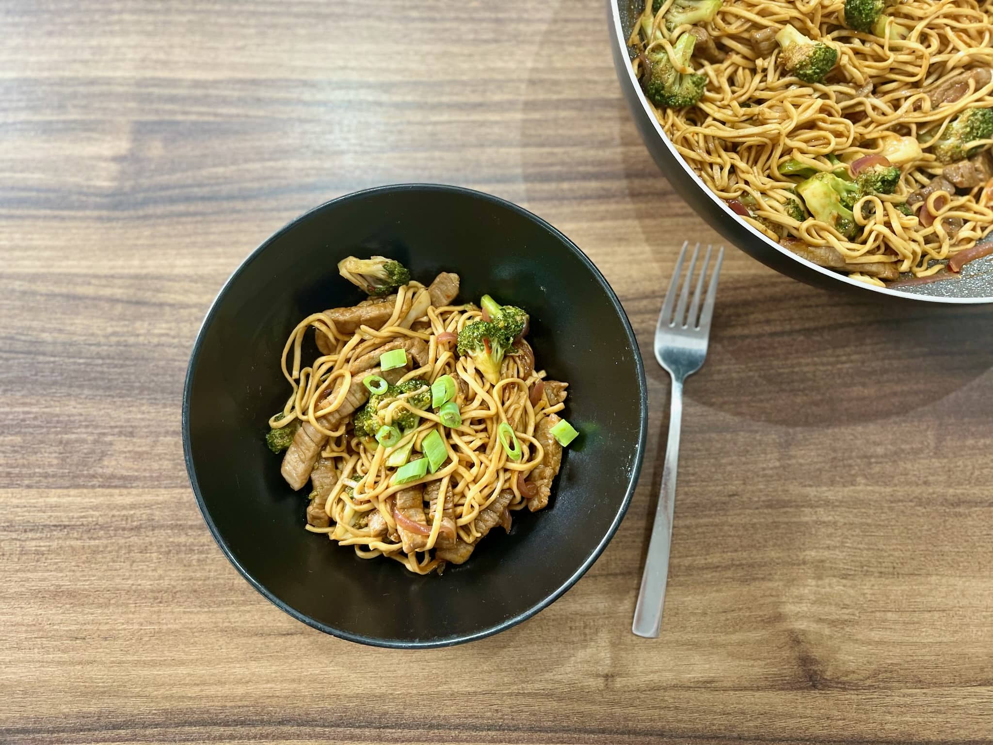 A portion of Beef Steak Stir Fry Noodles in a bowl on the tabletop with the rest in a pan on a side
