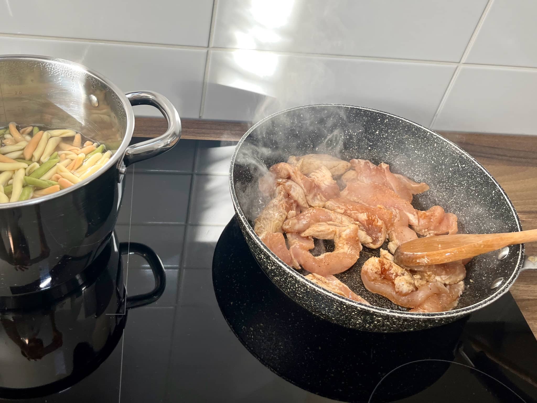 The pasta is cooking in a pan on the stovetop, and the chicken is frying in a large skillet over medium heat.