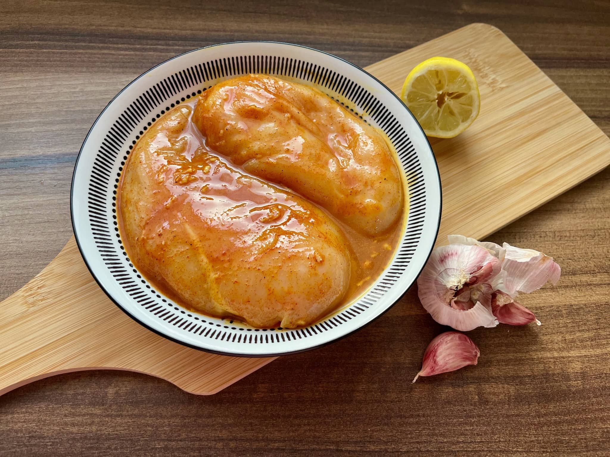 A bowl of marinated chicken breasts sits atop a chopping board, with lemon and garlic slices arranged on the side