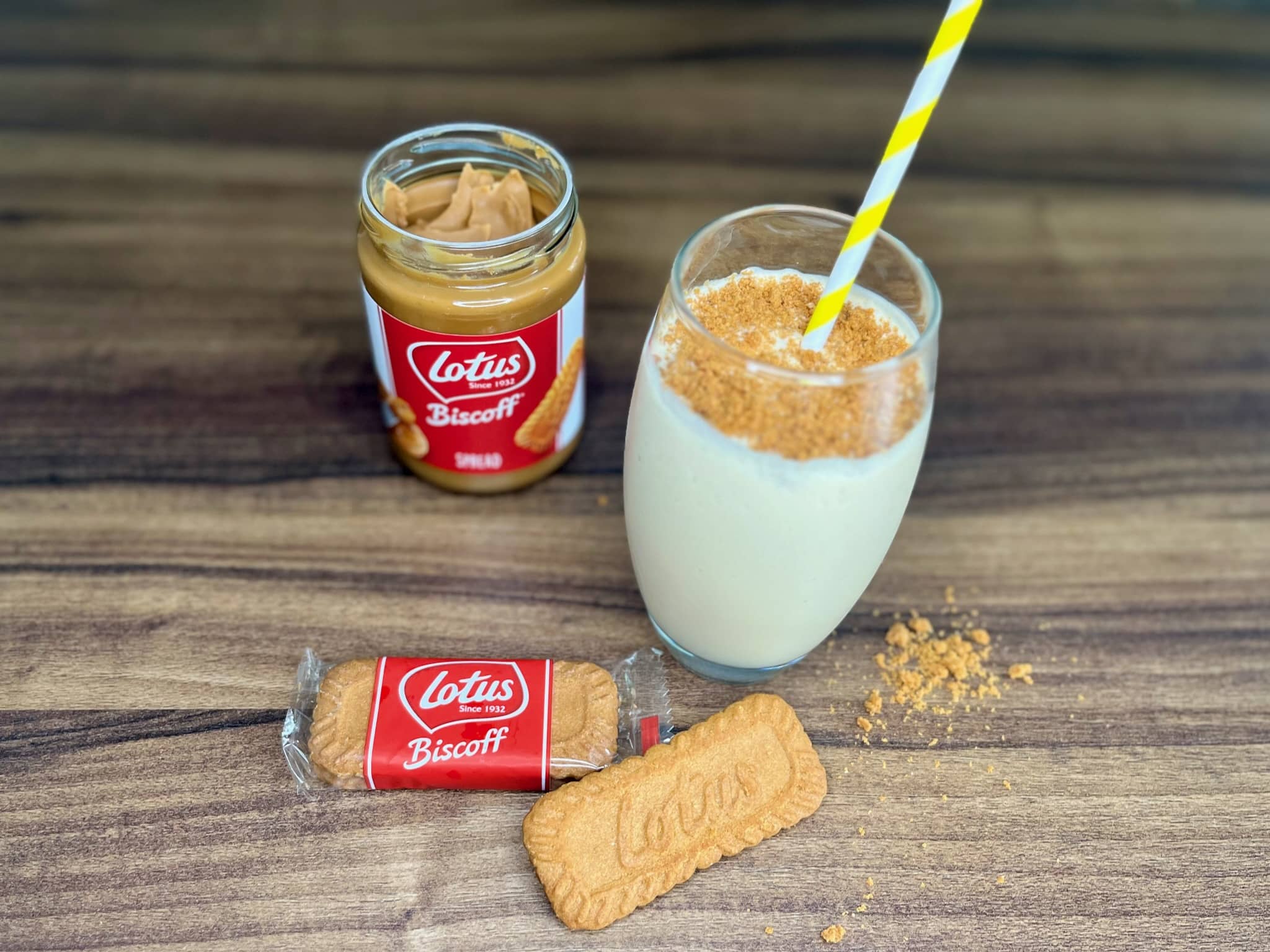 A tall glass of Biscoff milkshake with a straw, with Biscoff spread and biscuits on the side.
