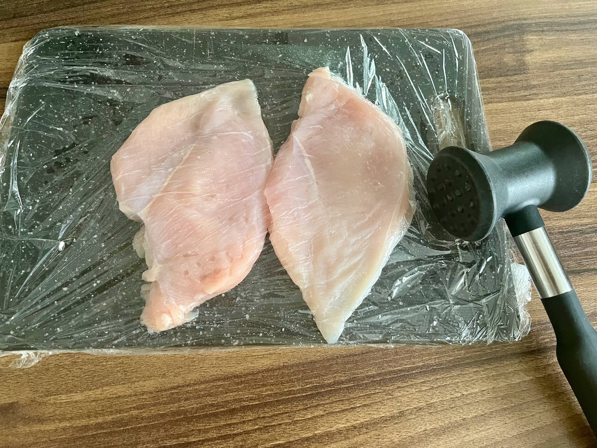 Chicken breasts on a chopping board, covered with cling film and levelled using a pestle