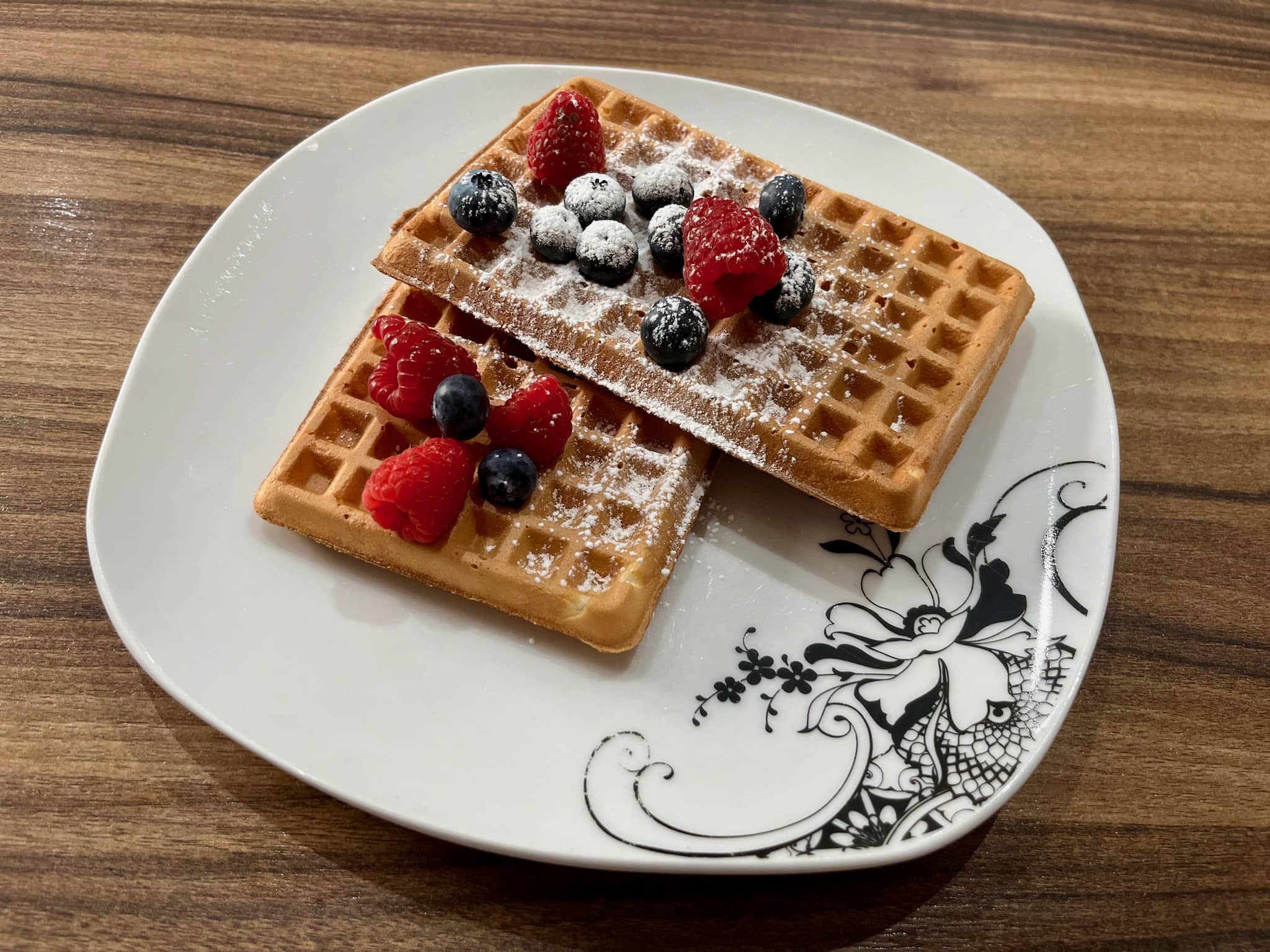 Two waffles on a plate decorated with icing sugar and some blueberries and raspberries