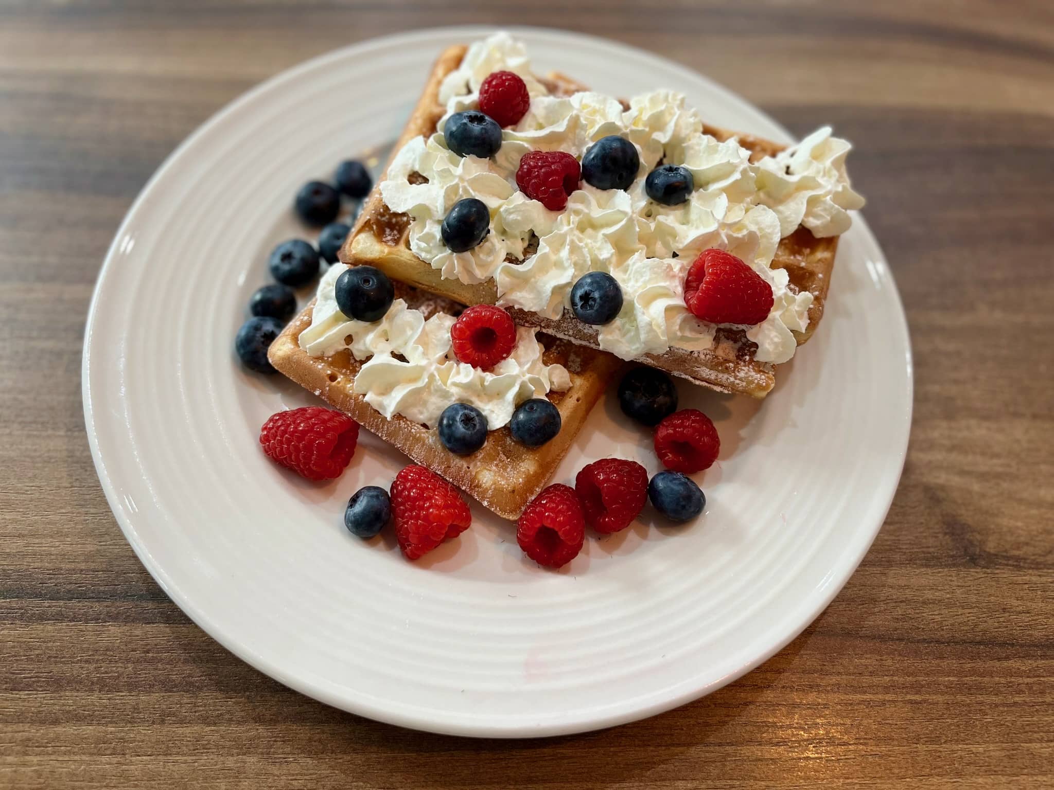 Two waffles on a plate decorated with whipped cream, icing sugar, blueberries and raspberries
