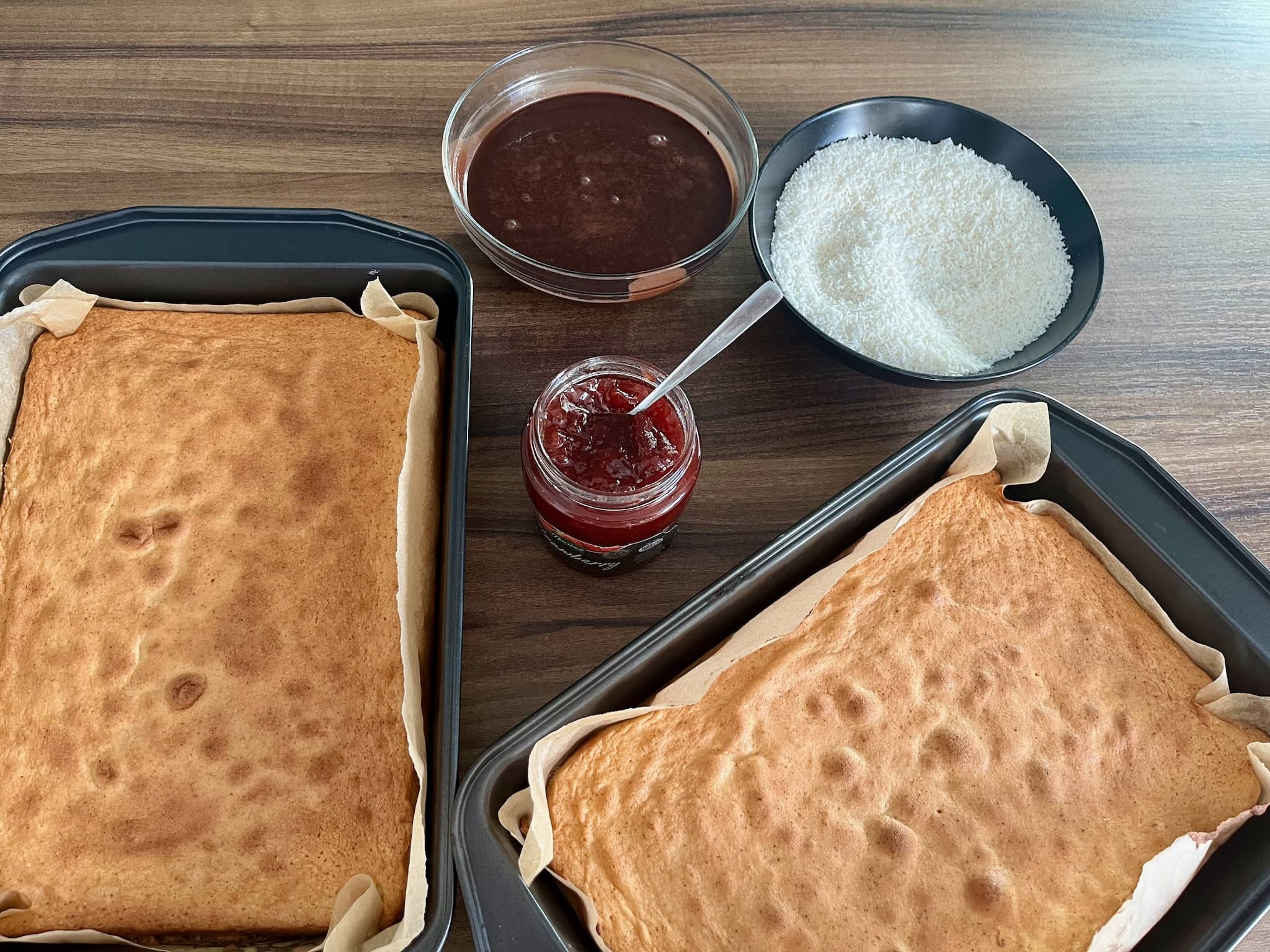 Two sponge cakes cooling down in trays with chocolate sauce and desiccated coconut in a bowl and strawberry jam in a jar