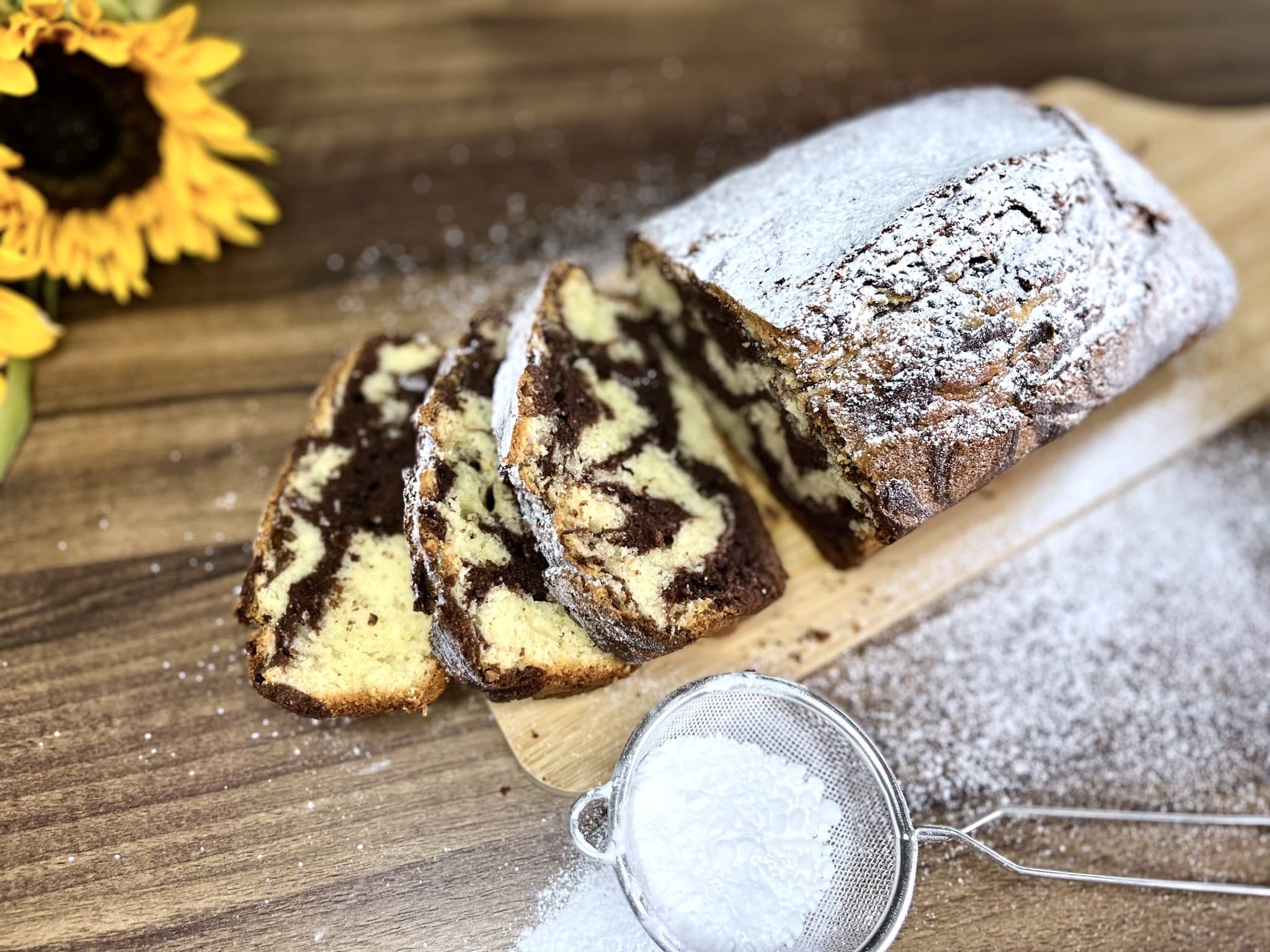 Marmurkowe ciasto na desce do krojenia, posypane cukrem pudrem, z dwoma ukrojonymi kawałkami z przodu