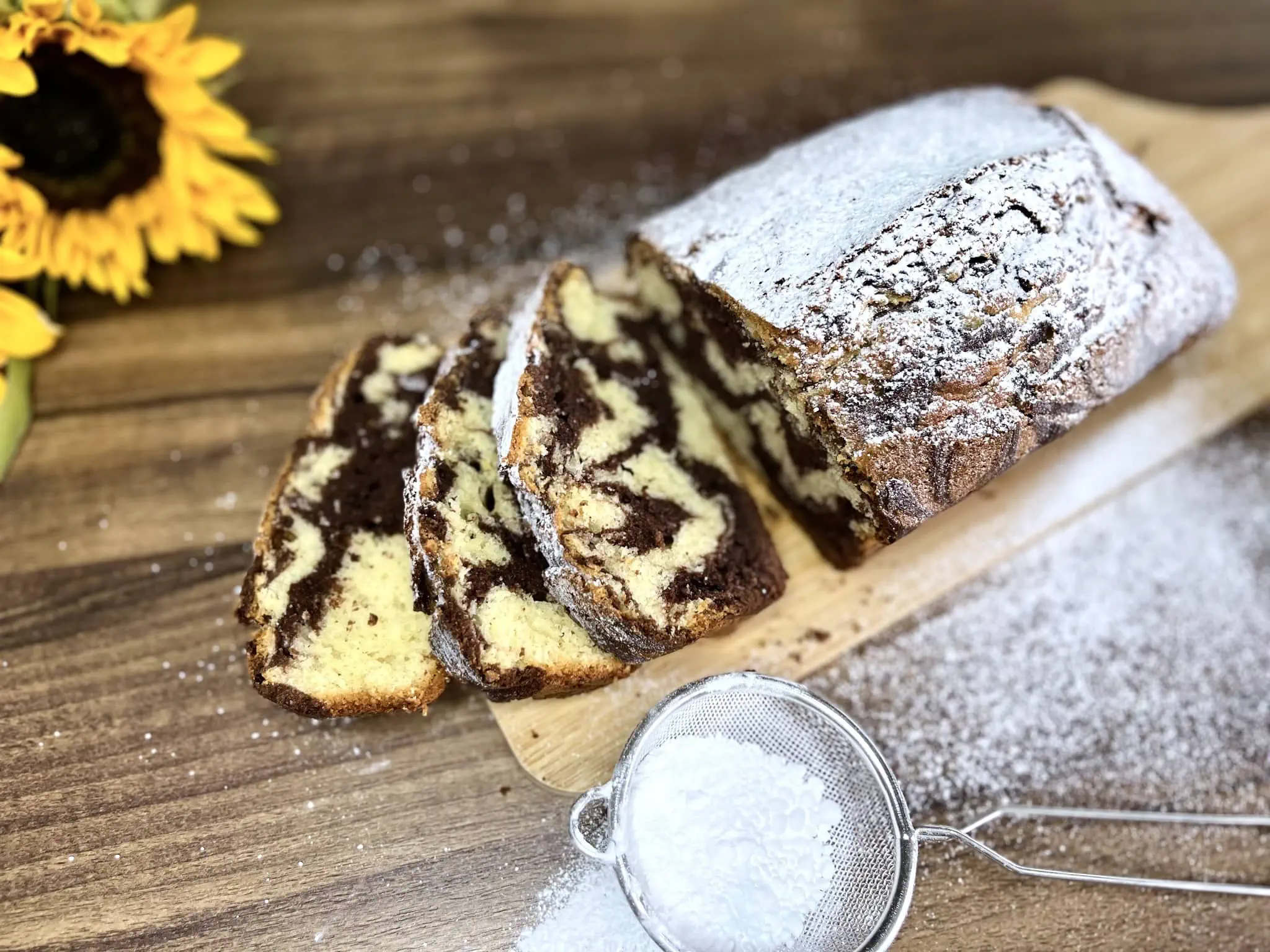 Marmurkowe ciasto na desce do krojenia, posypane cukrem pudrem, z dwoma ukrojonymi kawałkami z przodu