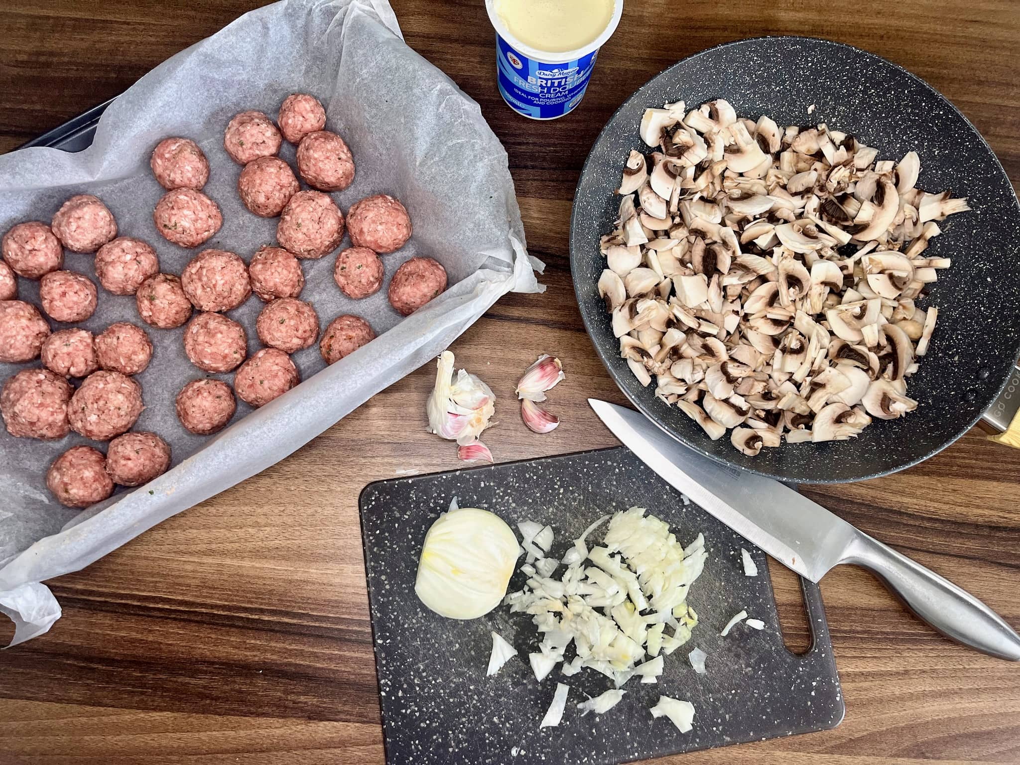 Meatballs in a tray, chopped onion on a chopping board, and sliced mushrooms in a pan