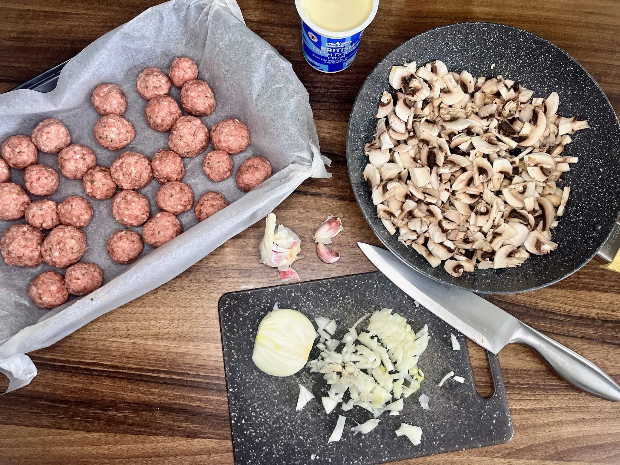 Klopsiki na blasze, posiekana cebula na desce i pokrojone pieczarki na patelni