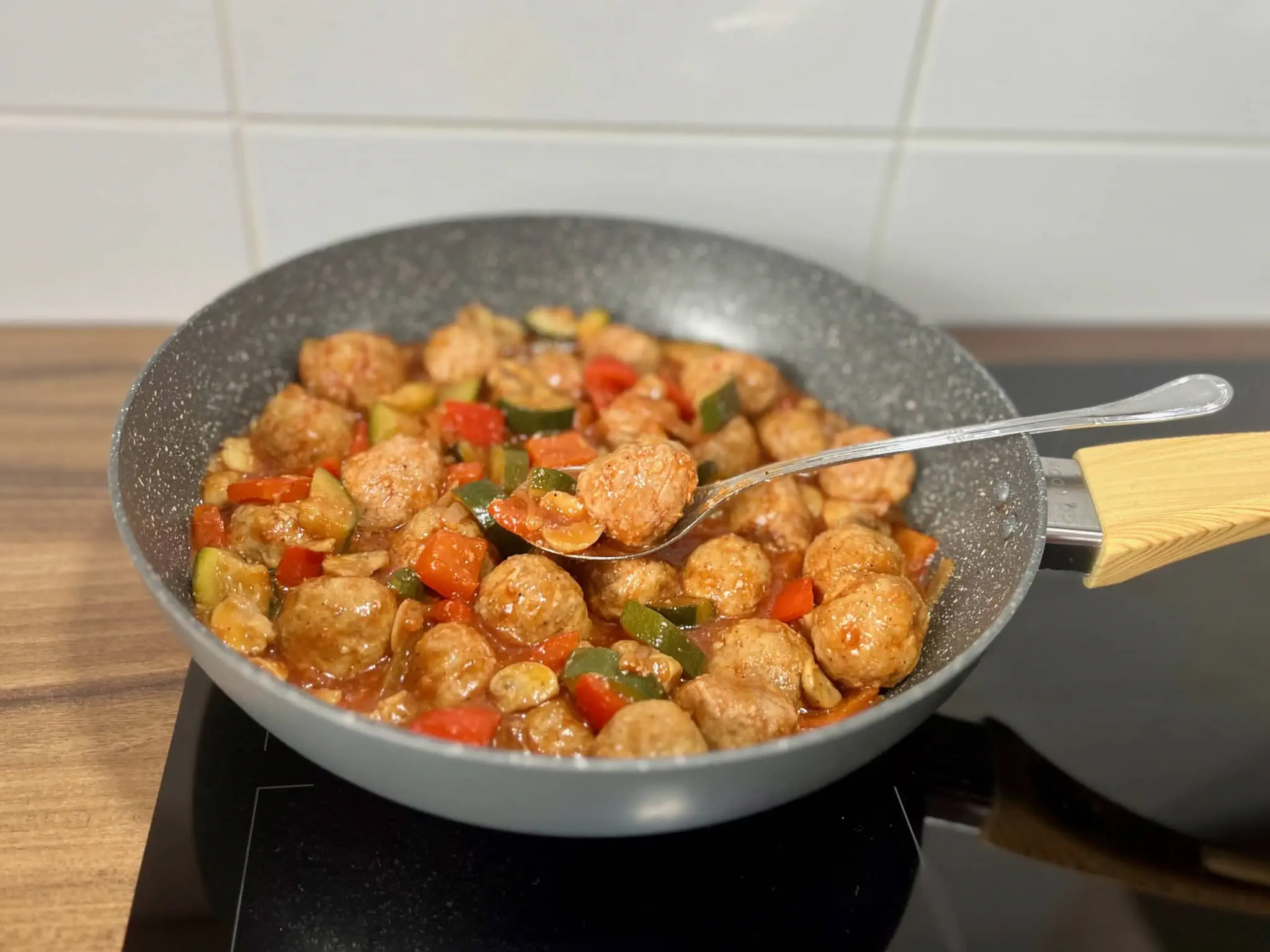 A spoon with a meatball on it, surrounded by meatballs and vegetables in a pan, all nicely covered in a sauce.
