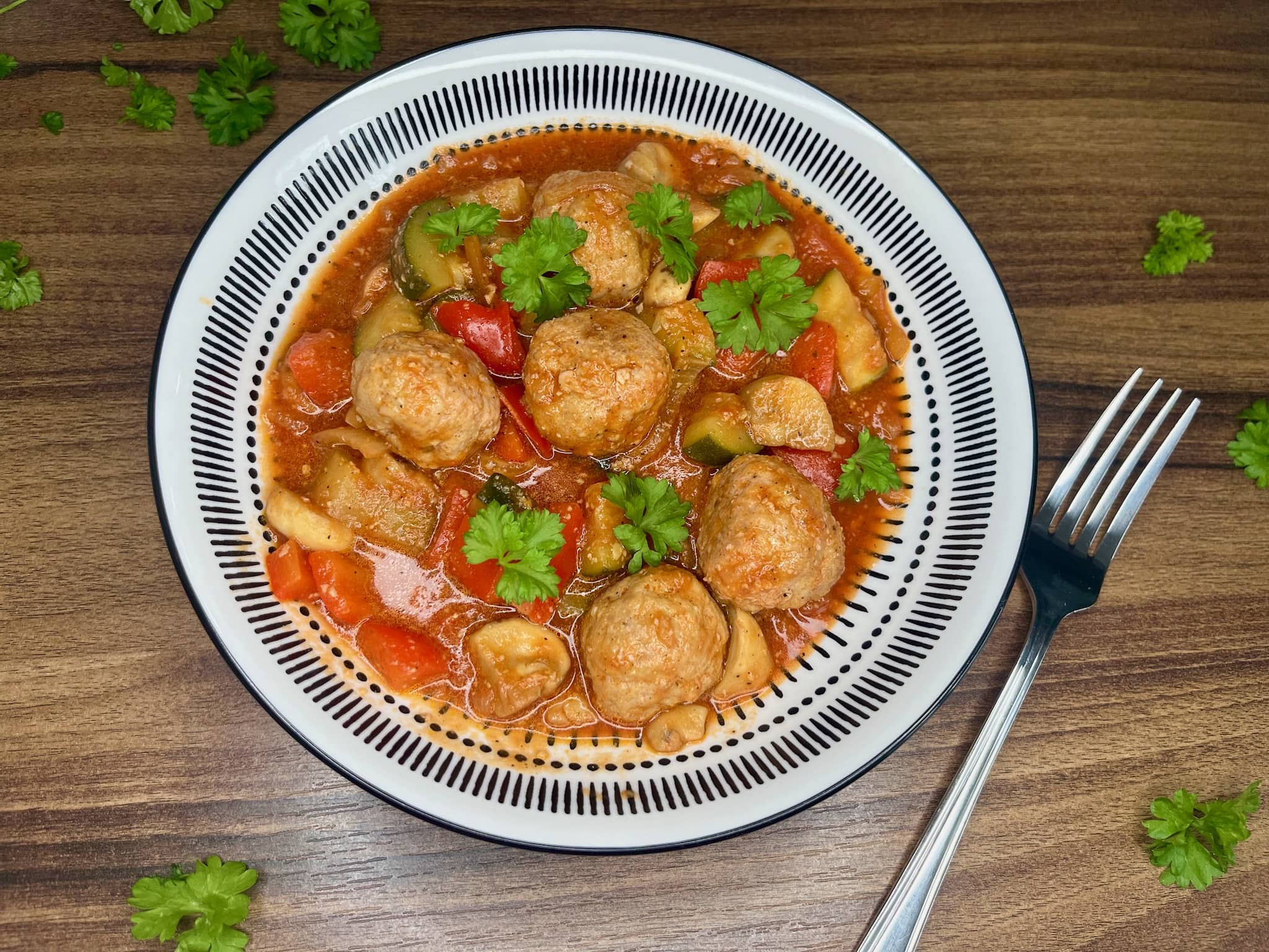 A bowl of meatballs in vegetable stew, surrounded by scattered parsley on a tabletop, is ready to be served.
