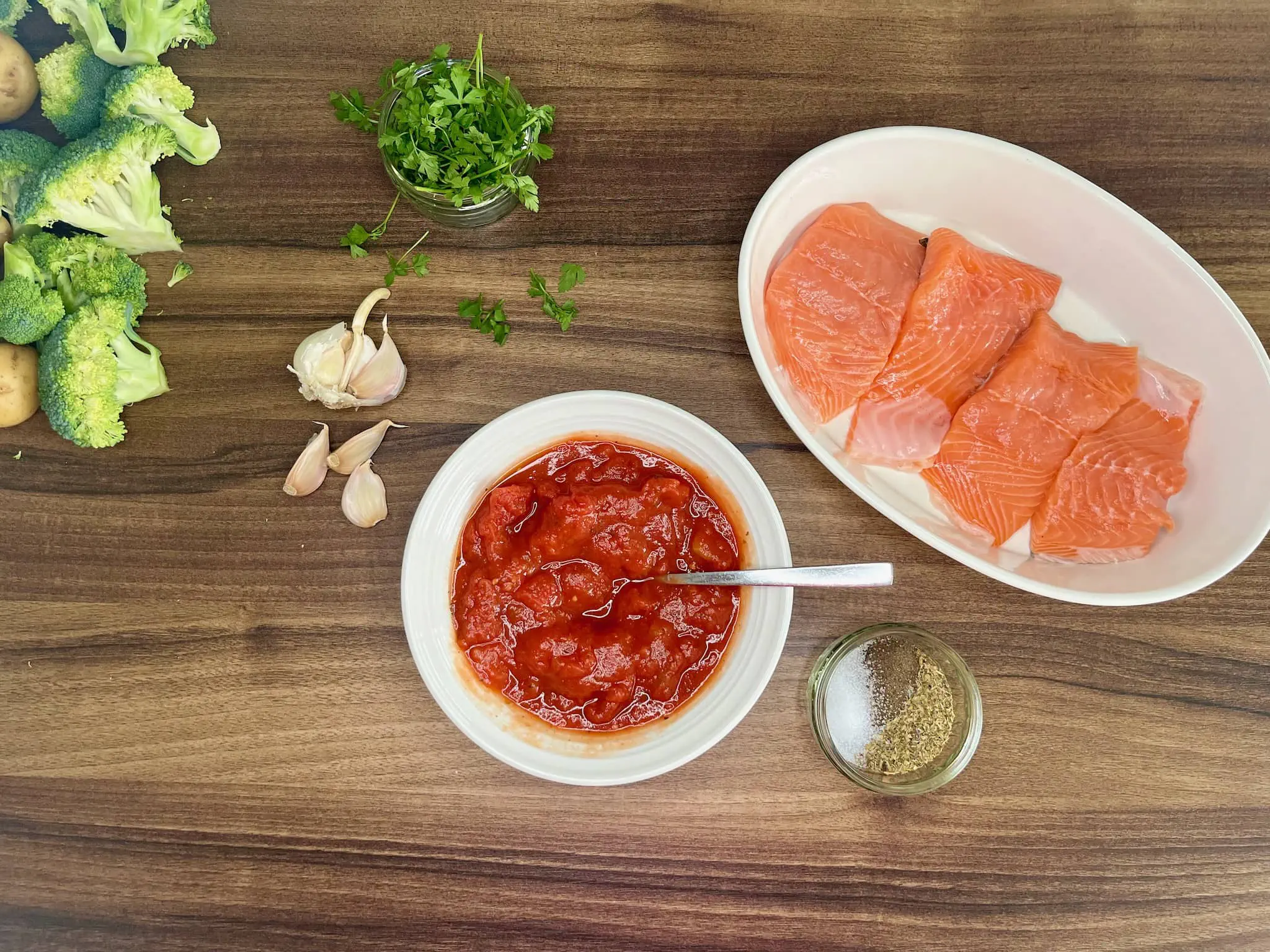 Pomidory w misce, przyprawy obok, a łosoś wyłożony w naczyniu żaroodpornym, gotowy do pieczenia w sosie pomidorowym