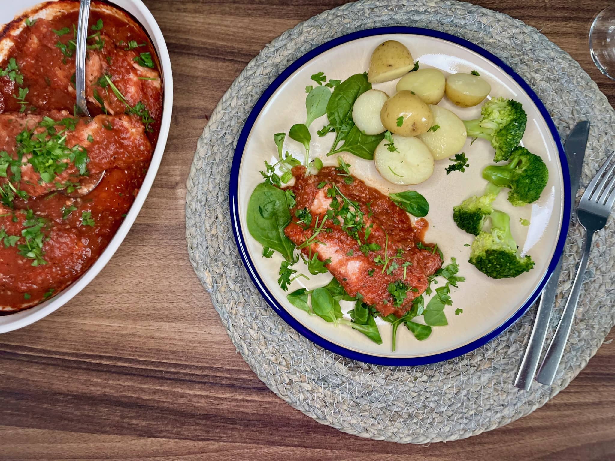 A plate with potion of oven-baked salmon in tomato sauce, with potatoes, broccoli, and a mixed salad with spinach