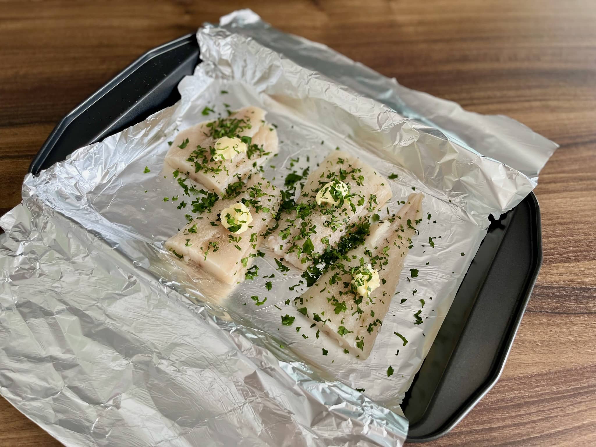 Fish fillets on a baking tray seasoned, with butter, garlic, parsley and lemon juice ready to bake.