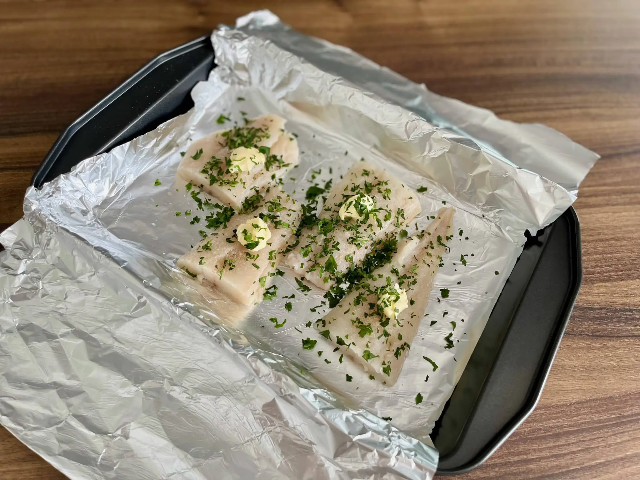 Filety rybne na blasze do pieczenia, przyprawione, z masłem, czosnkiem, pietruszką i sokiem z cytryny, gotowe do pieczenia.