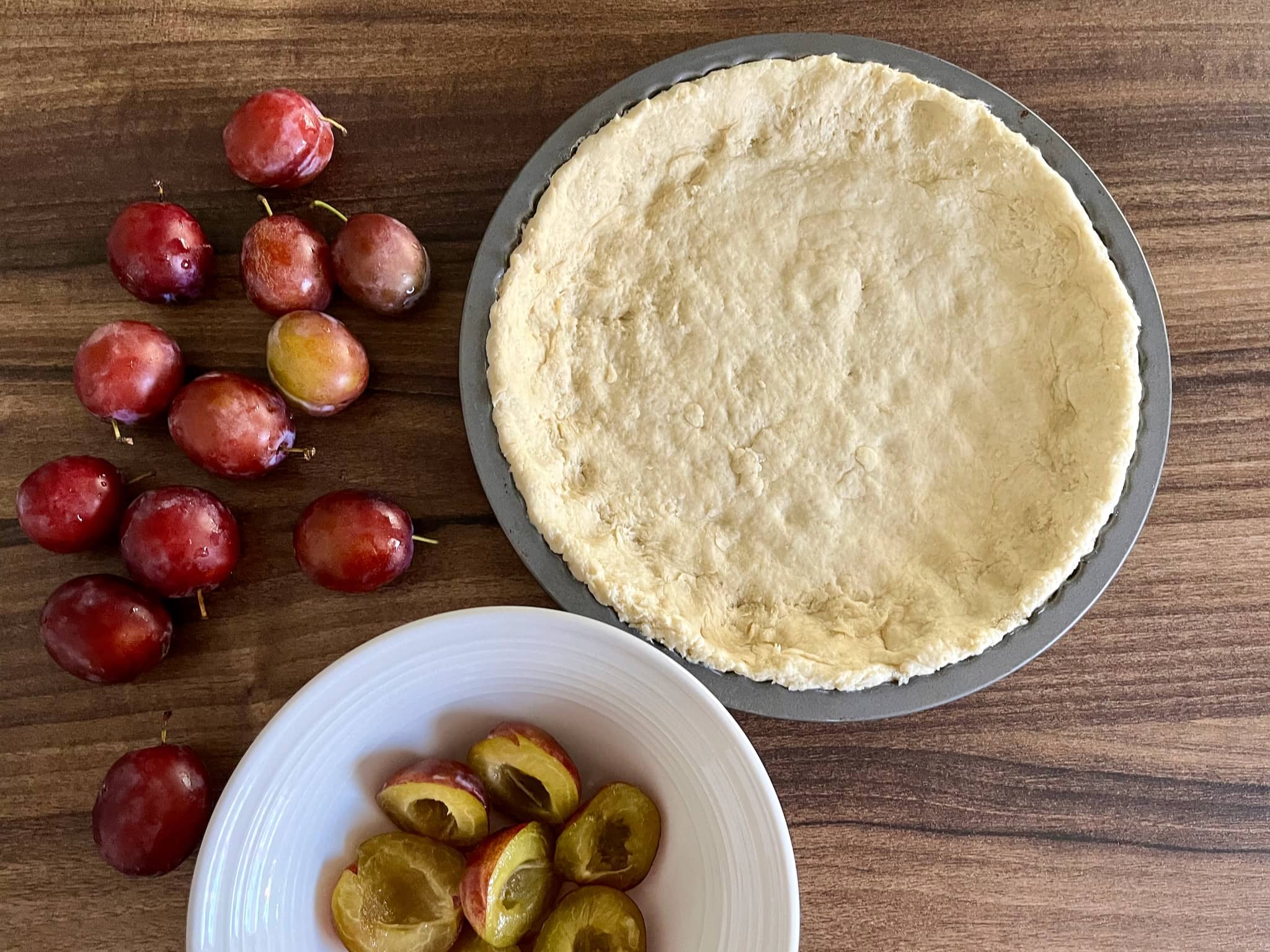 Dough in a tin with fresh plums on a side and halves of plums in a bowl