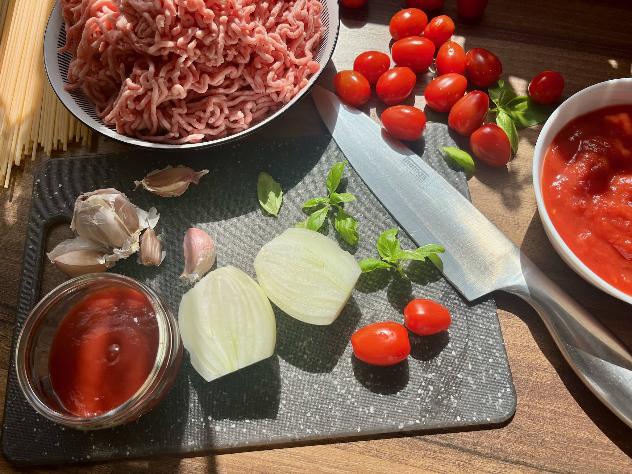 Minced meat and chopped tomatoes in a bowl, plum tomatoes, garlic and onion on a chopping board with ketchup on a side.