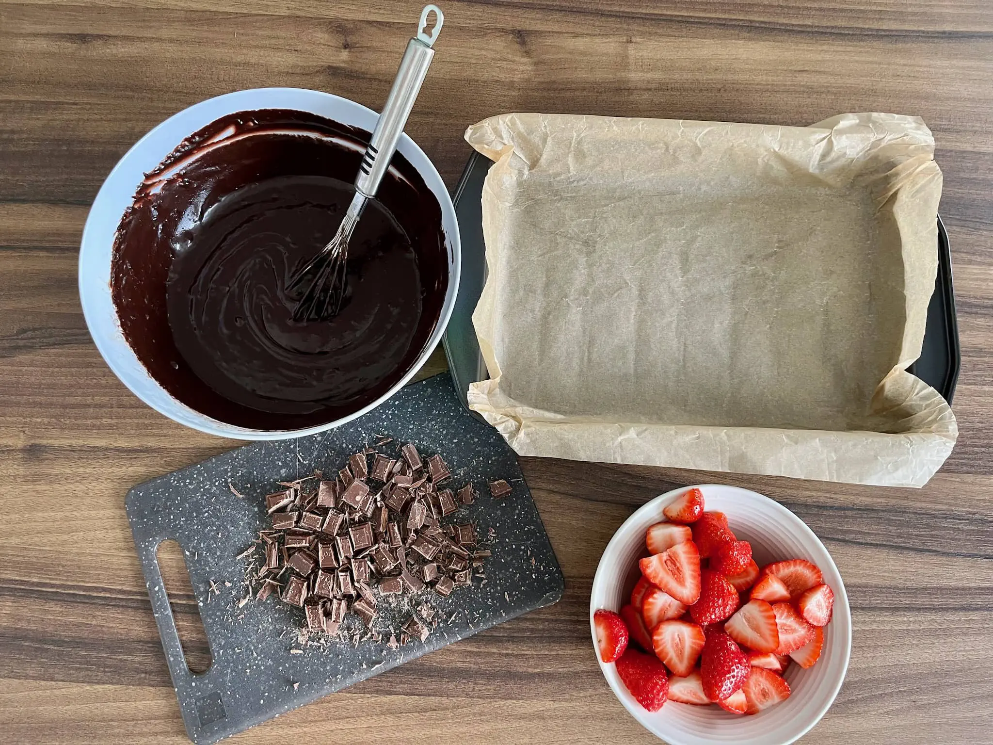 Masa na brownie w misce, posiekana czekolada, wyłożona blacha i przekrojone truskawki