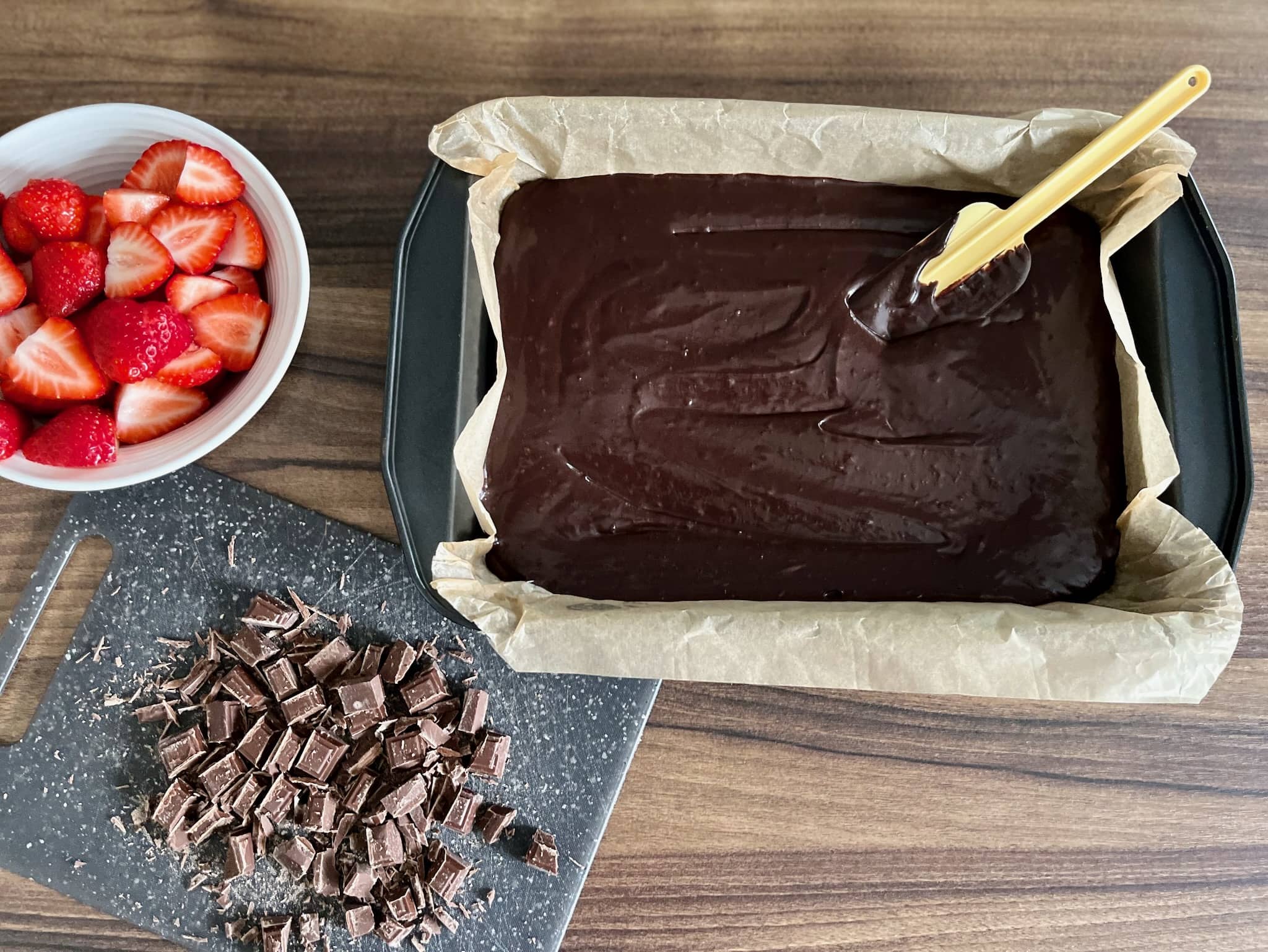 Brownie mix in a baking tray with strawberries and chocolate on a side still to add